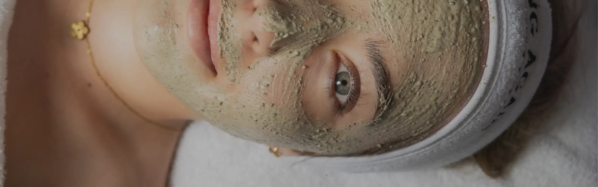A person wearing a white headband with black lettering and gold jewelry, their face covered in a textured greenish-brown facial mask.