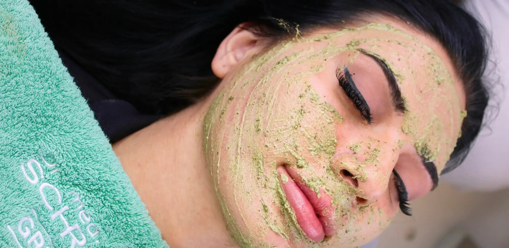 A woman with dark hair and long lashes has a greenish-brown facial mask applied to her face, resting on a teal towel.