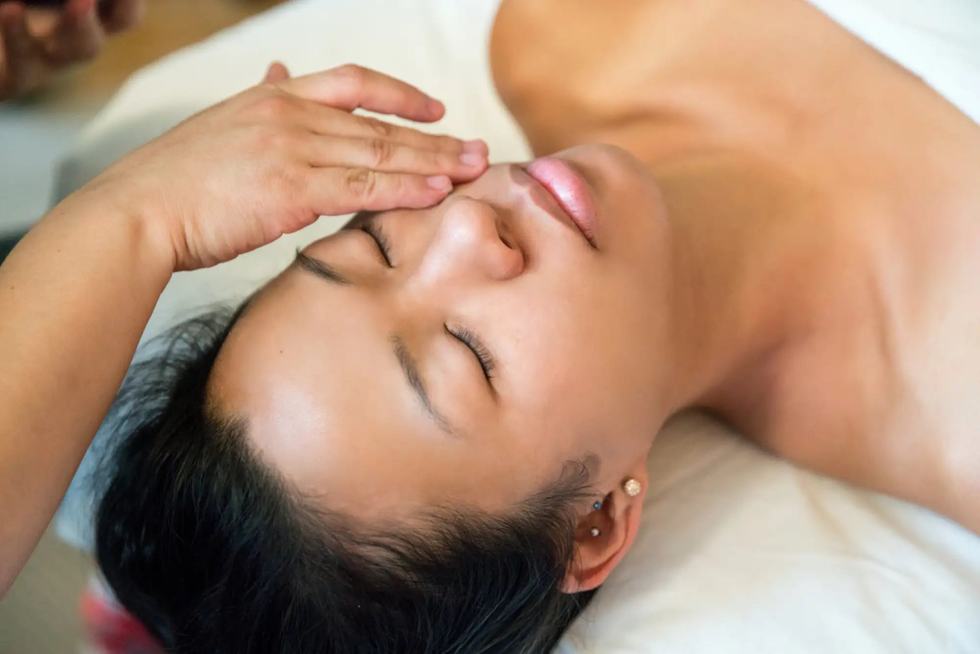 A woman with dark hair and a serene expression lies on her back, receiving a facial massage from another person’s hand gently resting near her forehead.